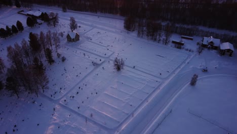 Aerial-view-during-winter-in-north-of-Sweden,-flying-over-a-road-with-cars-and-fields-covered-in-snow-slowly-tilting-up-revealing-a-frozen-lake-and-the-sun-setting-behind-the-mountains