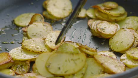 Close-Up-of-Crispy-Thinly-Sliced-Potatoes-Being-Cooked-Outside-in-Frying-Pan-and-Being-Gently-Turned-in-Slow-Motion-with-Herbs-de-Provence-and-Olive-Oil-4K