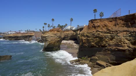 Sunset-Cliffs-Coastline,-San-Diego-California,-with-waves-crashing-agains-the-rocks-and-palm-trees-on-clear-day-with-blue-skies