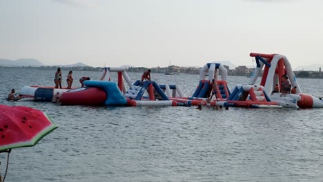 At-the-beach,-inflatable-slides-float-on-the-water,-filled-with-excited-children-and-adults-jumping,-sliding,-and-splashing-into-the-waves,-creating-joyful-chaos