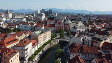 Aerial-drone-view-of-the-wonderful-city-of-Ljubljana-on-a-summer's-morning-with-city-and-mountains-in-background,-Tromostovje-bridge-and-Ljubljanica-river