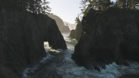 Aerial-view-of-waves-crashing-through-the-rocky-natural-bridges-along-the-Oregon-coast,-surrounded-by-forested-cliffs-and-rugged-terrain