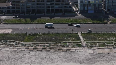 Aerial-drone-clip-of-a-single-cyclist-riding-past-pedestrians-along-a-beach-road-promenade-showing-the-sand-and-vegetation-on-the-beach-with-a-busy-road-and-apartments-in-the-background