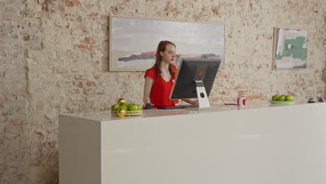 A-young-female-receptionist-stands-behind-a-large-desk-and-is-working-on-a-computer