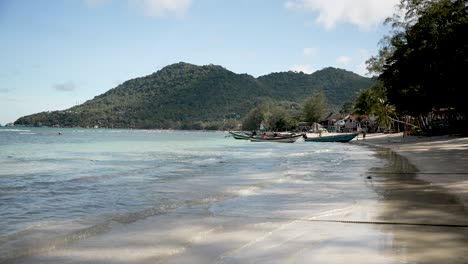 Eine-Ruhige-Tropische-Strandszene-Mit-Ruhigem-Türkisfarbenem-Wasser,-Am-Ufer-Vor-Anker-Liegenden-Booten-Und-üppigen-Grünen-Hügeln-Im-Hintergrund-Unter-Einem-Klaren-Blauen-Himmel