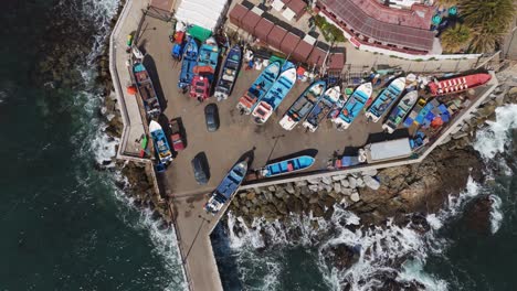 Toma-Aérea-De-Una-Variedad-De-Barcos-Amarrados-En-El-Puerto-De-Caleta-Quintay,-Mostrando-Una-Vibrante-Escena-Costera-Y-Las-Dinámicas-Olas-Del-Océano-Que-Rodean-El-Muelle.