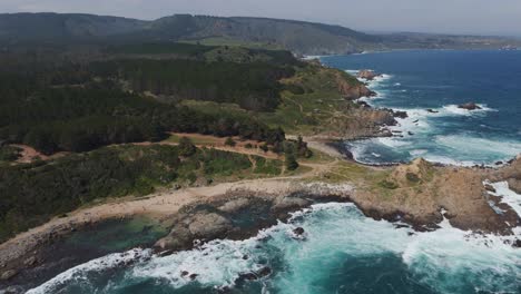 Türkisfarbene-Wellen-Brechen-An-Der-Felsigen-Küste-Von-Playa-Chica,-Chile,-Mit-üppigem-Grünen-Wald-Im-Hintergrund