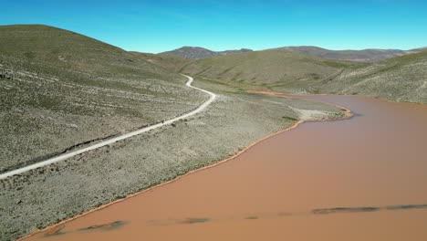 Aerial-view:-Moto-rides-dirt-road-past-muddy-lagoon-in-Peru-foothills