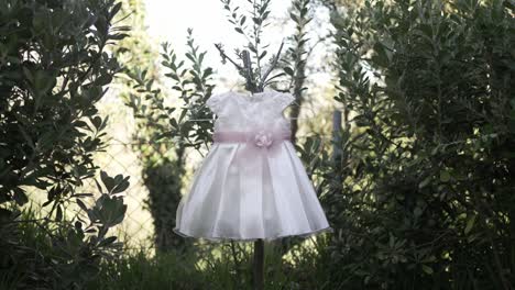 Baby-dress-with-floral-belt-hanging-between-green-bushes-outdoors,-soft-light