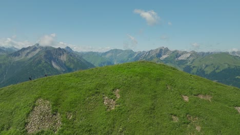 Two-hikers-trek-a-grassy-hilltop-with-a-vast-mountain-range-in-the-background-under-a-clear-sky