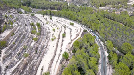 A-train-winds-through-a-green-forest-and-sandy-landscape-in-Calpe,-Spain,-aerial-view