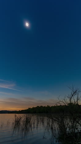 A-vertical-timelapse-video-of-a-total-solar-eclipse-recorded-from-start-to-finish,-with-the-darkened-sun-in-a-clear-daytime-sky-above-a-calm-lake