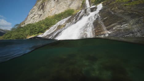 A-close-up-over-under-view-of-a-cascading-waterfall-flowing-into-the-calm-waters-of-Geiranger-Fjord,-surrounded-by-lush-greenery