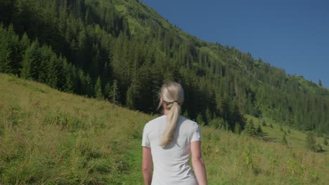 A-woman-hikes-through-a-green-mountain-meadow-under-a-clear-blue-sky-on-a-sunny-day