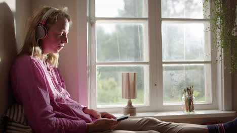 Woman-listens-to-music-with-headphones-basking-in-afternoon-window-sunlight