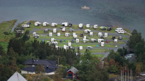 A-campsite-by-the-water-on-the-shores-of-the-Geiranger-fjord,-featuring-rows-of-RVs-and-tents-surrounded-by-lush-greenery