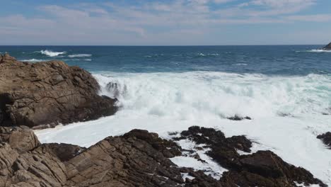 Aerial-view-of-powerful-ocean-waves-crashing-against-rugged-coastal-rocks,-under-a-bright-blue-sky,-symbolizing-the-untamed-beauty-and-strength-of-nature-at-its-finest