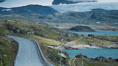 A-scenic-mountain-road-winds-through-a-rocky-landscape-dotted-with-lakes,-cabins-scattered-throughout-the-valley,-and-a-glacier-dominating-the-background