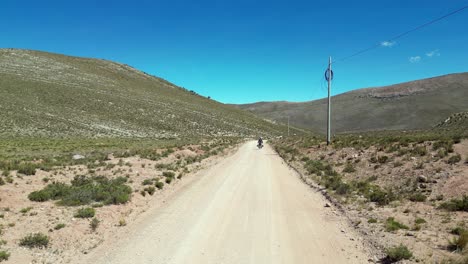 Motorcycle-rides-from-high-angle-camera-on-arid-foothills-dirt-road