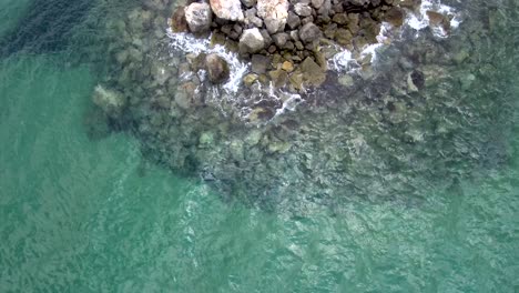 Aerial-view-of-stunning-sea-turquoise-in-the-summer-with-rocks-that-water-pops-up-on-them-in-the-area-of-Agia-Paraskevi-Halkidiki,-Greece,-up-and-side-movement-by-drone