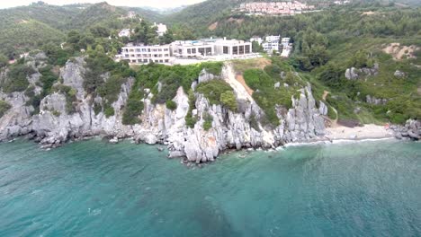 Aerial-view-of-the-small-turquoise-beach-with-rocks,-under-a-hot-Springs-building,-in-the-area-of-Agia-Paraskevi,-Halkidiki,-circular-movement-by-drone