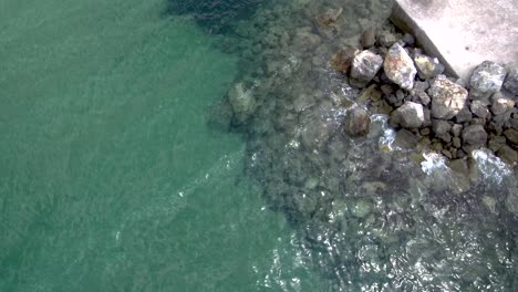 Aerial-view-of-stunning-turquoise-sea-in-the-summer-with-pier-and-rocks-that-water-pops-up-on-them-in-the-area-of-Agia-Paraskevi-Halkidiki,-Greece,-up-and-side-movement-by-drone