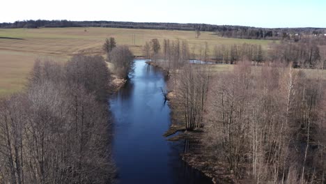 Drohnenschuss-Flussabwärts-Eines-Mystischen-Schwarzen-Flusses-In-Schweden-Mit-Feldern-Und-Wald-Im-Hintergrund