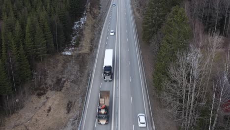 Drone-shot-of-highway-in-Sweden-running-through-forest,-panning-upwards-to-sky-revealing-forest,-trucks-and-cars-driving-by
