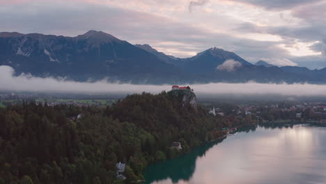 Aerial-shot-of-Bled-Castle-on-top-of-a-mountain-with-low-lying-clouds-over-a-lake