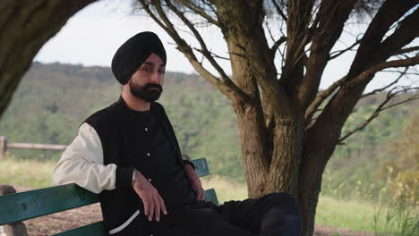 Hübscher-Punjabi-Sikh-Mann,-Der-Auf-Einer-Bank-Mit-Naturhintergrund-Sitzt
