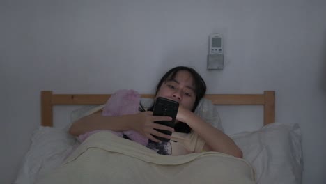 Southeast-asian-woman-sits-in-bed-yawns-while-scrolling-on-phone,-dozes-off-to-sleep