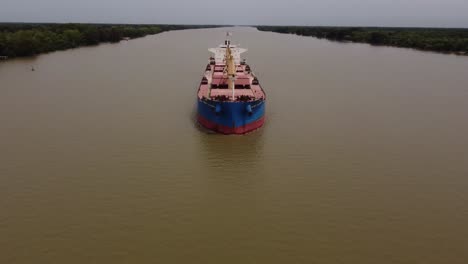 Aerial-ascending-shot-of-industrial-Bulk-Carrier-Cargo-Ship-sailing-on-Parana-River-beside-Rainforest---Tilt-down