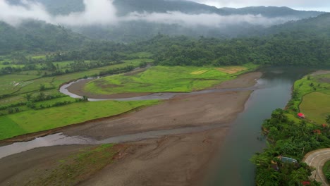 Slow-aerial-dolly,-panoramic-view-of-mud-flats-by-farmlands-and-river-banks,-cloudy-mountains-behind
