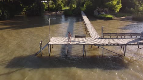 Aerial-orbiting-shot-of-man-in-bathing-trunks-standing-on-wooden-dock-in-front-of-Parana-River-during-sunny-day---Argentina,-South-America