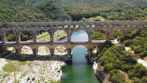 Tilted-Aerial-View-of-the-Pont-du-Gard-Aqueduct-over-the-Hérault-River,-with-a-Canoe-in-the-Background-on-a-Summer-Day-in-Occitanie