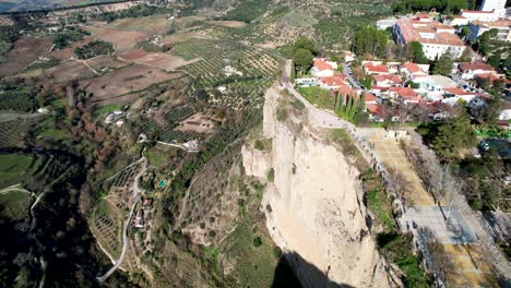 Drone-clip-slowly-moving-towards-terracotta-builidings-on-top-of-rock-cliff,-with-agricultural-land-on-the-slopes-below,-in-Ronda,-Spain