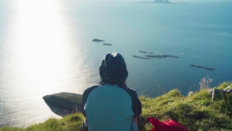 Rear-Of-A-Person-In-A-Hoody-Sitting-On-A-The-Mountain-Edge-In-Lovund-Island,-Norway