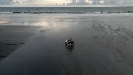 Aerial:-Man-rides-motorcycle-through-shallow-water-on-wide-sand-beach
