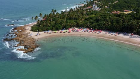 Rotating-aerial-drone-shot-of-the-popular-tropical-Coquerinhos-beach-surrounded-by-palm-trees-and-covered-in-umbrellas-with-tourists-swimming-in-a-natural-pool-in-Conde,-Paraiba,-Brazil
