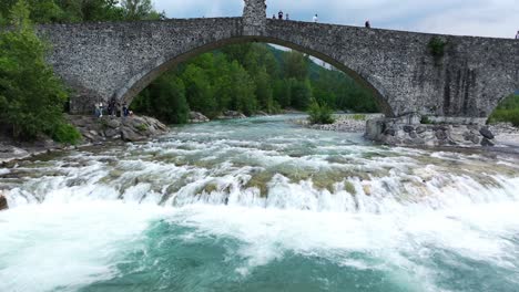 Drone-flying-under-Gobbo-bridge-along-Trebbia-river,-Bobbio-in-Italy