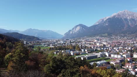 Vista-Aérea-De-La-Ciudad-De-Innsbruck-Con-Sus-Edificios-Desde-Las-Colinas-De-Un-Bosque-En-Un-Soleado-Día-De-Otoño-Y-Un-Cielo-Azul-Y-Al-Fondo-Los-Alpes-Con-Sus-Picos,-Tirol,-Austria,-Europa