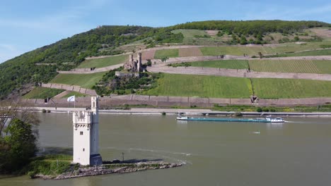 Freighter-cargo-ship-barge-navigating-down-Rhine-River-cruising-past-toll-tower-and-Ehrenfels-Castle,-Germany