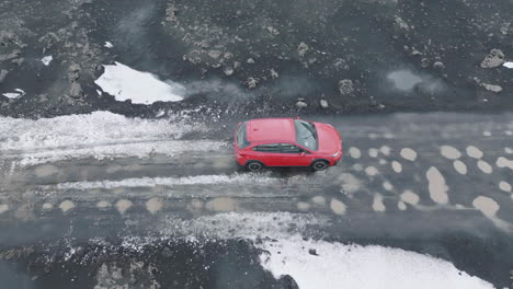 Red-Car-Moving-on-Wet-Road-Surrounded-With-Snow,-Ice-and-Lava-Fields-in-Landscape-of-Iceland,-Drone-Shot