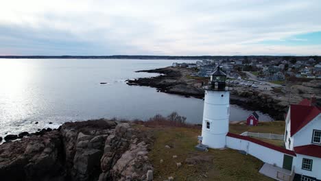 Lighthouse-on-a-rocky-island-and-a-quaint-village-on-the-water-during-dusk