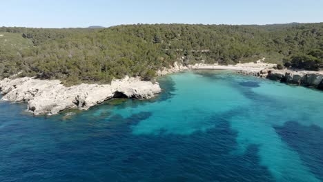 Aerial-view-of-Cala-Escorxada,-the-virgin-beach-in-Menorca,-Spain-with-blue-water-and-a-hill-nearby-with-green-trees-and-blue-clear-sky-in-the-background