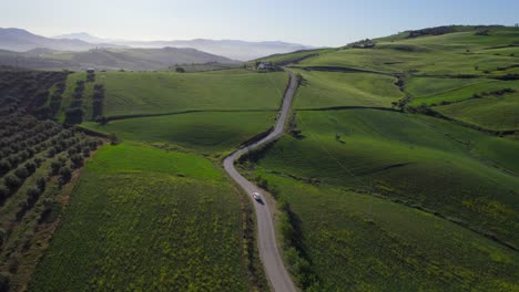 Drone-Siguiendo-Una-Camioneta-Blanca-Conduciendo-A-Lo-Largo-De-Un-Camino-Estrecho-En-El-Campo-Con-Grandes-Colinas-Verdes