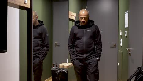 Bald-Young-Indian-Male-Pacing-Past-Large-Mirror-In-Hallway-Talking-Wirelessly-Dressed-In-Black-Trousers-and-Hoody