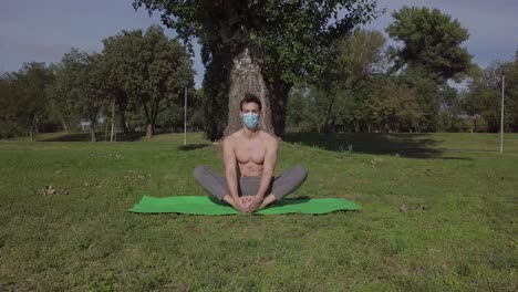 Attractive-man-with-mask-practicing-yoga-simple-pose-exercise-on-the-mat-in-the-park