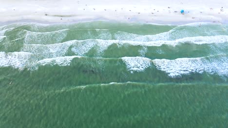 Birdseye-drone-clip-of-green-water-waves-rolling-onto-white-sand-beach,-with-people-sunbathing-below