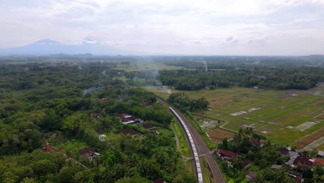 Aerial-view-of-riding-train-in-rural-area-with-forest-trees-and-mountains-in-background---Indonesia,Asia